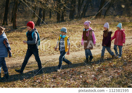 Active little classmates, boys and girls, in warm hats and jackets enjoying nature walk on clear day. Outdoor activities for children's development. Active little classmates, boys and girls, in warm hats and jackets enjoying nature walk on clear day. Outdoor activities for children's development. 112381595