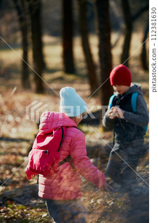 Little kids, classmates dressed in layered clothing exploring wooded area on sunny day on environmental lesson. Outdoor activities for children's development. Little kids, classmates dressed in layered clothing exploring wooded area on sunny day on environmental lesson. Outdoor activities for children's development. 112381630