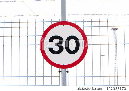 Speed limit sign attached to wire mesh fence. Maximum thirty kilometers per hour. Safety on road background. White round sign red border line. Concept of following traffic rules. Symbol Slow down turn Speed limit sign attached to wire mesh fence. Maximum thirty kilometers per hour. Safety on road background. White round sign red border line. Concept of following traffic rules. Symbol Slow down turn 112382170