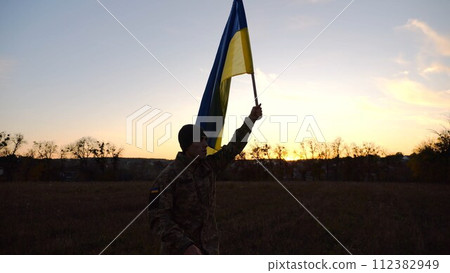 Soldier of ukrainian army going with raised blue-yellow banner on field at sunset. Young male military in uniform walks with flag of Ukraine at meadow. Concept of victory against russian aggression 112382949