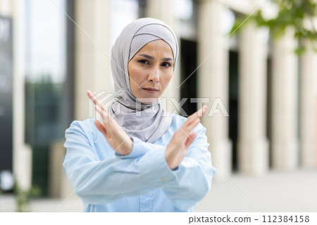 Empowered adult Muslim woman wearing a hijab crosses her arms in an 'X' gesture, signaling stop or denial outside. 112384158