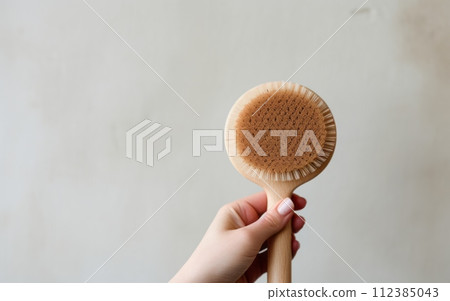A hand holding a circular, wooden-bristle dry brush against a neutral background 112385043