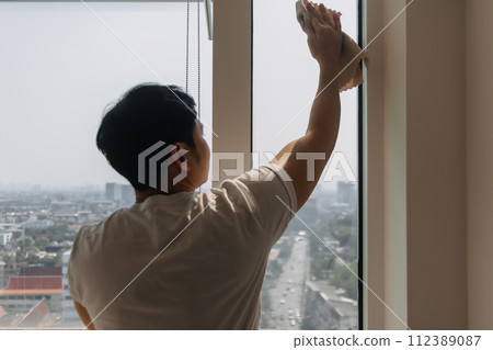Backside view of asian Thai man wiping window glass in room apartment with city view, keep glass sparking clean, housework concept. Backside view of asian Thai man wiping window glass in room apartment with city view, keep glass sparking clean, housework concept. 112389087