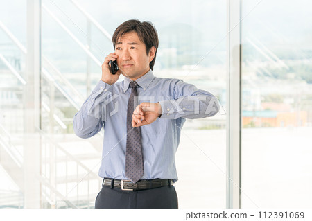 中年男性亞裔商人在辦公大樓用智慧型手機交談（遲到、麻煩、麻煩、會議） 112391069