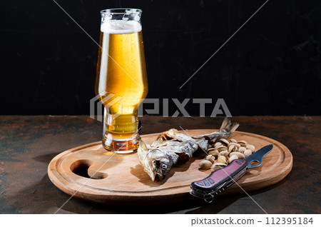 Light beer in a glass and dried fish. Beer and snack on the kitchen board. Fish and knife on the table. 112395184