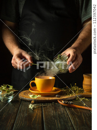 A man brews tea with medicinal herbs. Traditional medicine concept at home for treating influenza virus or cold. Adding dry mint to a mug 112397042