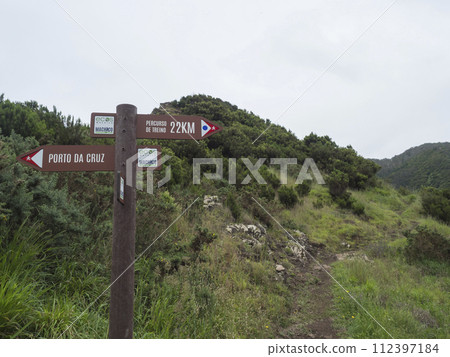 Green hills and meadow with footpath and tourist signpost pointing at Porto da Cruz in one direction and Percurso de treino at Machico in other. Green hills and meadow with footpath and tourist signpost pointing at Porto da Cruz in one direction and Percurso de treino at Machico in other. 112397184