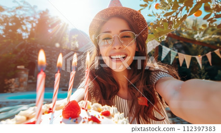 Playful young woman takes a selfie with a birthday cake with one candle. Sunny daylight. 112397383