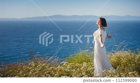 Beautiful Asian young woman in white dress outdoor in flower field under Rhodes city and above sea during sunset. embracing fresh air and engaging in outdoor activities. strong wind - element of 112399080