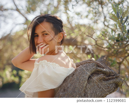 Beautiful Asian young woman in white dress outdoor near olive tree. embracing fresh air and engaging in outdoor activities. Friluftsliv concept means spending as much time outdoors as possible Beautiful Asian young woman in white dress outdoor near olive tree. embracing fresh air and engaging in outdoor activities. Friluftsliv concept means spending as much time outdoors as possible 112399081
