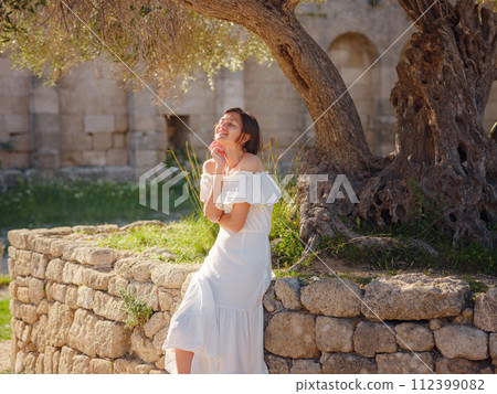 Beautiful Asian young woman in white dress outdoor. Acropolis of Rhodes Famous ruins of ancient settlement with various buildings such as stadium and theater. 112399082