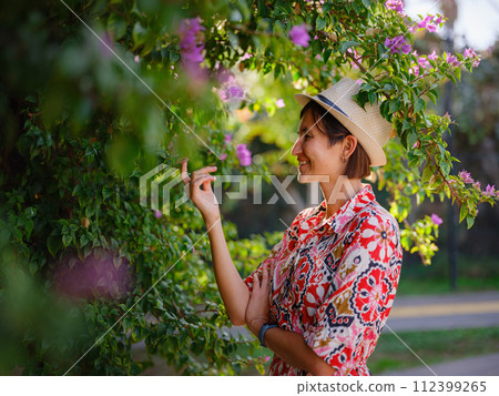 young asian woman stands in sunny green park, admiring blooming bougainvillea bush. The flower's bright colors are beautiful contrast to park's lush greenery. 112399265