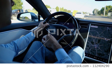 Male hand of businessman holds steering wheel while driving electric car at city. Businessperson operating electrical vehicle on urban road. Young man commuting to work at sunny day. Close up Male hand of businessman holds steering wheel while driving electric car at city. Businessperson operating electrical vehicle on urban road. Young man commuting to work at sunny day. Close up 112404290