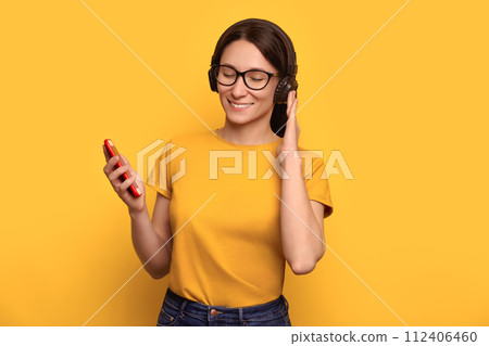 Melomaniac girl listening to music with wireless headphones on yellow studio wall 112406460