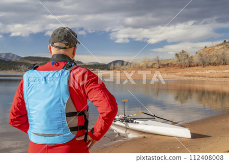 senior man wearing drysuit and life jacket with a rowing shell on lake shore, winter in northern Colorado 112408008