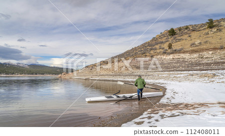 Senior male rower with a coastal rowing shell and hatchet oars on a shore of Carter Lake in fall or winter scenery in northern Colorado. Senior male rower with a coastal rowing shell and hatchet oars on a shore of Carter Lake in fall or winter scenery in northern Colorado. 112408011