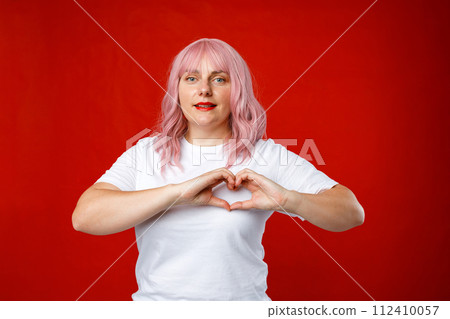 Attractive 30s brunette young woman feels happy and romantic shapes heart gesture expresses tender feelings poses against red background. Emotional face. Human emotions, facial expression concept Attractive 30s brunette young woman feels happy and romantic shapes heart gesture expresses tender feelings poses against red background. Emotional face. Human emotions, facial expression concept 112410057