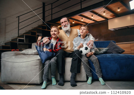 Likable concentrated senior bearded grandfather with his cheerful 10-15s grandsons spending their free time at revision the basketball game on tv 112410920