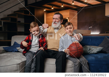 Concentrated senior bearded grandpa with his happy grandsons spending their leisure at revision the basketball championship on tv at home. 112410922