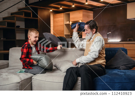 Happy boy with funny modern grandpa fighting with pillows having fun together sitting on comfortable couch at home. 112410923