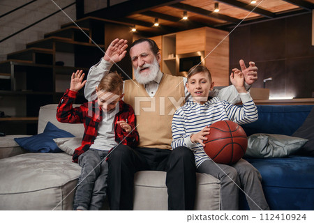 Concentrated senior bearded grandpa with his happy grandsons spending their leisure at revision the basketball championship on tv at home. 112410924