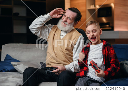 Cute little boy with grandfather sitting on sofa and playing video game with game pad 112410940