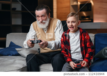Cute little boy with grandfather sitting on sofa and playing video game with game pad 112410944
