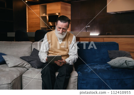 Elderly man learns tablet computer. Electronic gadgets for the senior people. A man with a stylish beard using tablet for web surfing. Elderly man learns tablet computer. Electronic gadgets for the senior people. A man with a stylish beard using tablet for web surfing. 112410955