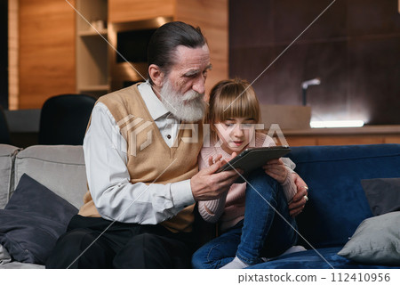 Grandpa with his granddaughter using tablet pc at cozy home. Little girl teach her granddad to use smart devices. 112410956