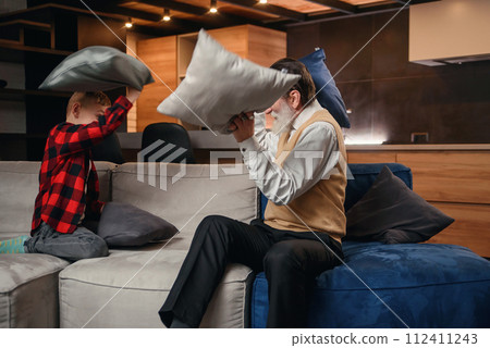 Happy boy with funny modern grandpa fighting with pillows having fun together sitting on comfortable couch at home. Happy boy with funny modern grandpa fighting with pillows having fun together sitting on comfortable couch at home. 112411243