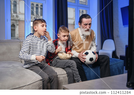 Likable concentrated senior bearded grandfather with his cheerful 10-15s grandsons spending their free time at revision the basketball game on tv Likable concentrated senior bearded grandfather with his cheerful 10-15s grandsons spending their free time at revision the basketball game on tv 112411284