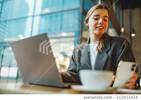 Woman manager reading message on phone and work on laptop in cafe. Distance work concept Woman manager reading message on phone and work on laptop in cafe. Distance work concept 112411414
