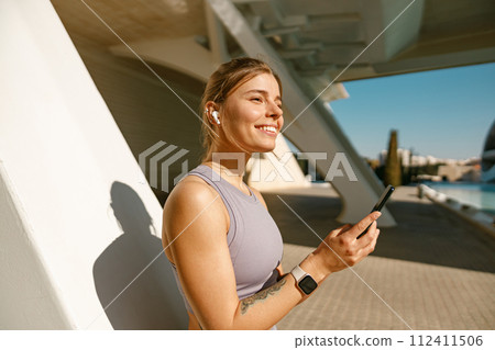 A young woman in sportswear is resting after a workout outside while standing with smartphone A young woman in sportswear is resting after a workout outside while standing with smartphone 112411506