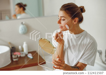 Young woman massaging face with jade roller and looking at round cosmetic mirror in bathroom 112411507