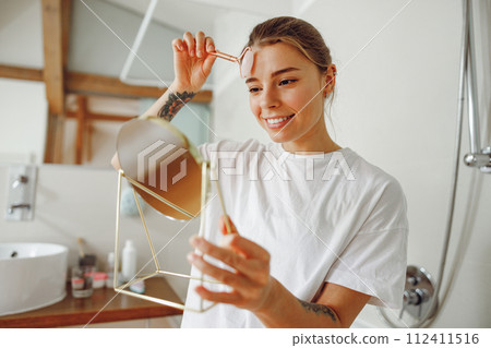 Smiling woman massaging her face with jade roller massager and looking at round cosmetic mirror Smiling woman massaging her face with jade roller massager and looking at round cosmetic mirror 112411516