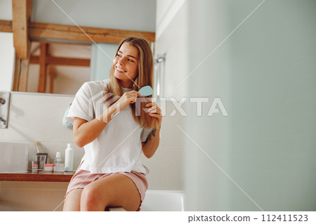 Smiling young woman brushing her hair with comb while standing in bathroom near mirror 112411523
