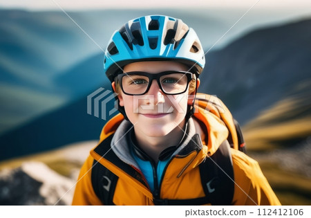 Young boy wearing helmet and glasses stands confidently before towering mountain backdrop ready for adventure and exploration. He may be gearing up for bicycle ride or some other outdoor activity. Young boy wearing helmet and glasses stands confidently before towering mountain backdrop ready for adventure and exploration. He may be gearing up for bicycle ride or some other outdoor activity. 112412106