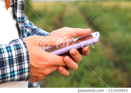 A close-up shot captures a male Caucasian farmer's hands firmly grasping a smartphone in an outdoor setting A close-up shot captures a male Caucasian farmer's hands firmly grasping a smartphone in an outdoor setting 112420953