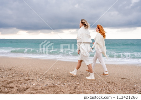 Women sea walk friendship spring. Two girlfriends, redhead and blonde, middle-aged walk along the sandy beach of the sea, dressed in white clothes. Against the backdrop of a cloudy sky and the winter Women sea walk friendship spring. Two girlfriends, redhead and blonde, middle-aged walk along the sandy beach of the sea, dressed in white clothes. Against the backdrop of a cloudy sky and the winter 112421266