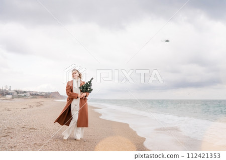 Blond woman Christmas tree sea. Christmas portrait of a happy woman walking along the beach and holding a Christmas tree in her hands. She is wearing a brown coat and a white suit. 112421353