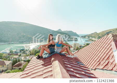 Two women sitting on a red roof, enjoying the view of the town and the sea. Rooftop vantage point. In the background, there are several boats visible on the water, adding to the picturesque scene. Two women sitting on a red roof, enjoying the view of the town and the sea. Rooftop vantage point. In the background, there are several boats visible on the water, adding to the picturesque scene. 112421366