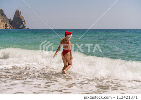 A woman in Santa hat on the seashore, dressed in a red swimsuit. New Year's celebration in a hot country A woman in Santa hat on the seashore, dressed in a red swimsuit. New Year's celebration in a hot country 112421371