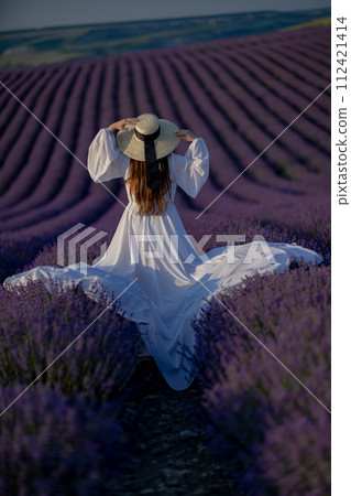 Back view woman lavender sunset. Happy woman in white dress holds lavender bouquet. Aromatherapy concept, lavender oil, photo session in lavender Back view woman lavender sunset. Happy woman in white dress holds lavender bouquet. Aromatherapy concept, lavender oil, photo session in lavender 112421414