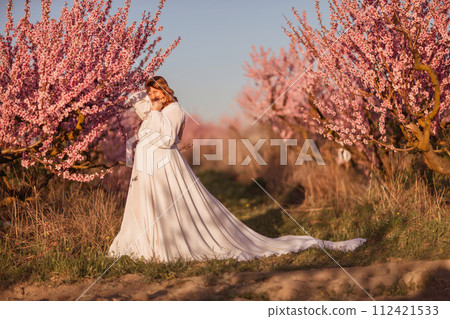Woman blooming peach orchard. Against the backdrop of a picturesque peach orchard, a woman in a long white dress enjoys a peaceful walk in the park, surrounded by the beauty of nature. Woman blooming peach orchard. Against the backdrop of a picturesque peach orchard, a woman in a long white dress enjoys a peaceful walk in the park, surrounded by the beauty of nature. 112421533