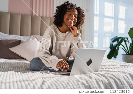 African American girl using laptop in bed at home office typing chatting reading writing email. Young black woman having virtual meeting online chat video call conference. Work learning from home African American girl using laptop in bed at home office typing chatting reading writing email. Young black woman having virtual meeting online chat video call conference. Work learning from home 112423915