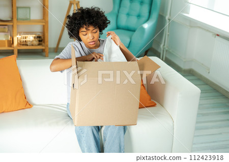 African girl unpacking delivery looking in box. Happy woman opening carton box. Female getting parcel looking at delivered goods items. Satisfied client positive feedback quick express deliver African girl unpacking delivery looking in box. Happy woman opening carton box. Female getting parcel looking at delivered goods items. Satisfied client positive feedback quick express deliver 112423918