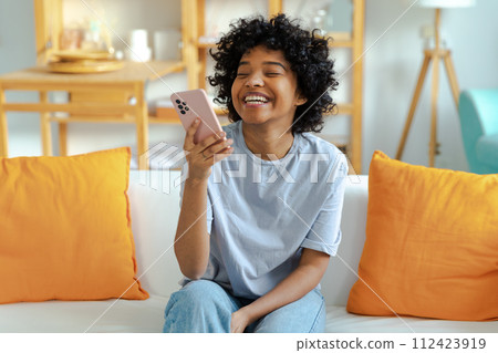 Excited happy young black african american woman holding cell phone laughing feeling joy getting mobile message. Overjoyed girl laughing aloud sitting on couch watching funny video reading news Excited happy young black african american woman holding cell phone laughing feeling joy getting mobile message. Overjoyed girl laughing aloud sitting on couch watching funny video reading news 112423919