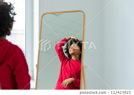 Love yourself. Beautiful young smiling african american woman dancing touching curly hair enjoying her mirror reflection. Black lady looking at mirror looking confident and happy. Self love concept Love yourself. Beautiful young smiling african american woman dancing touching curly hair enjoying her mirror reflection. Black lady looking at mirror looking confident and happy. Self love concept 112423923