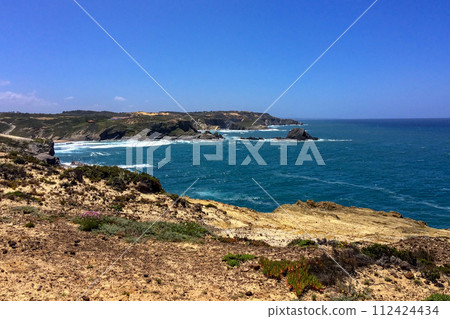 Ocean waves hit the jagged cliffs on a sunny day. 112424434