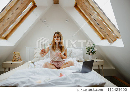 Woman is sitting on a bed with a laptop and a cell phone in a cozy house with houseplants 112425600